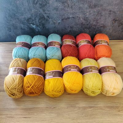 12 balls of yarn in shades of blue, red, orange, yellow and brown laid out on a wooden surface.