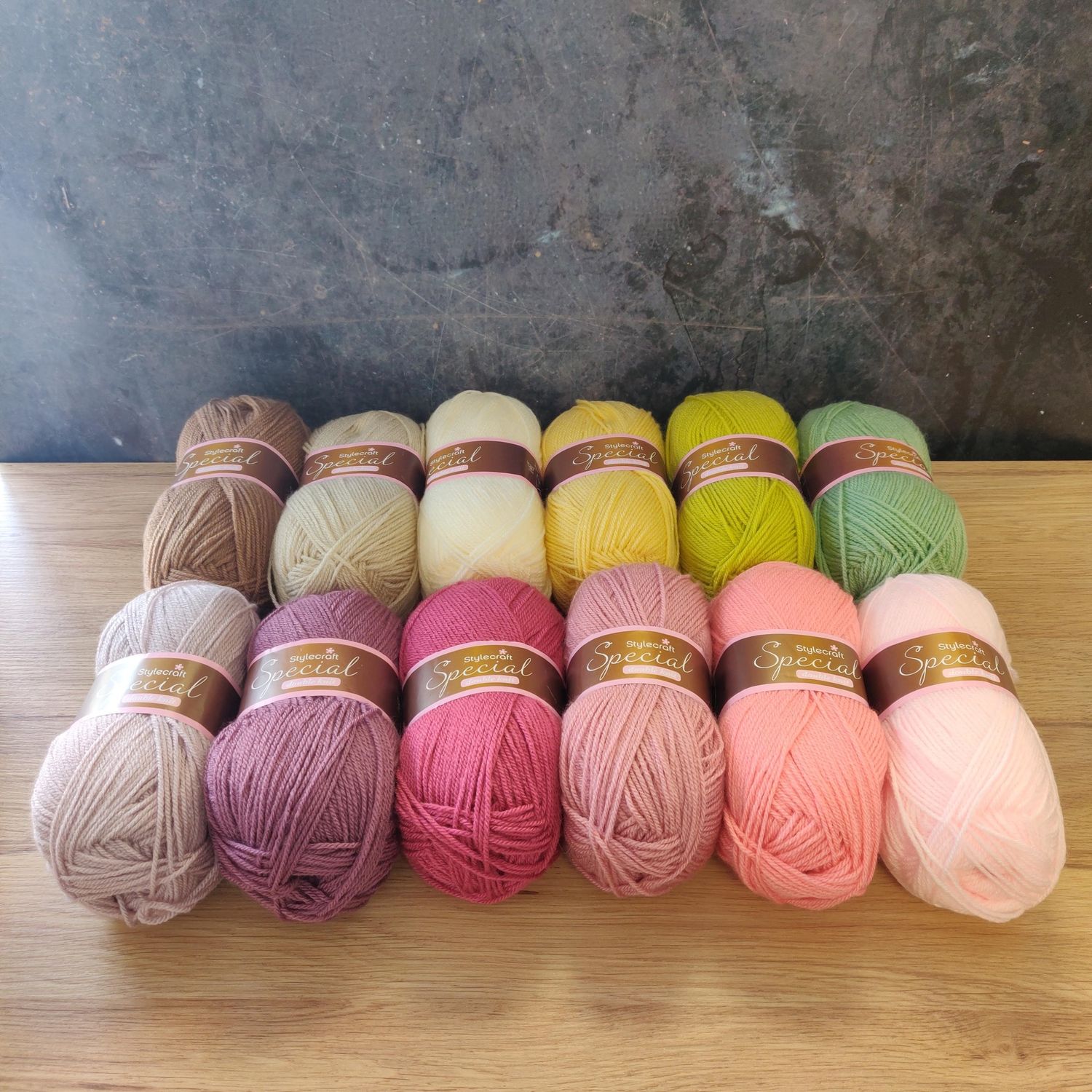 12 balls of yarn in shades of brown, cream, green, purple and pink laid out on a wooden surface.