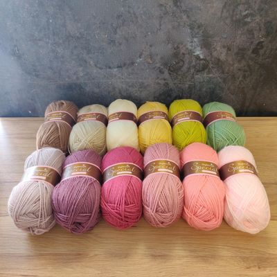 12 balls of yarn in shades of brown, cream, green, purple and pink laid out on a wooden surface.