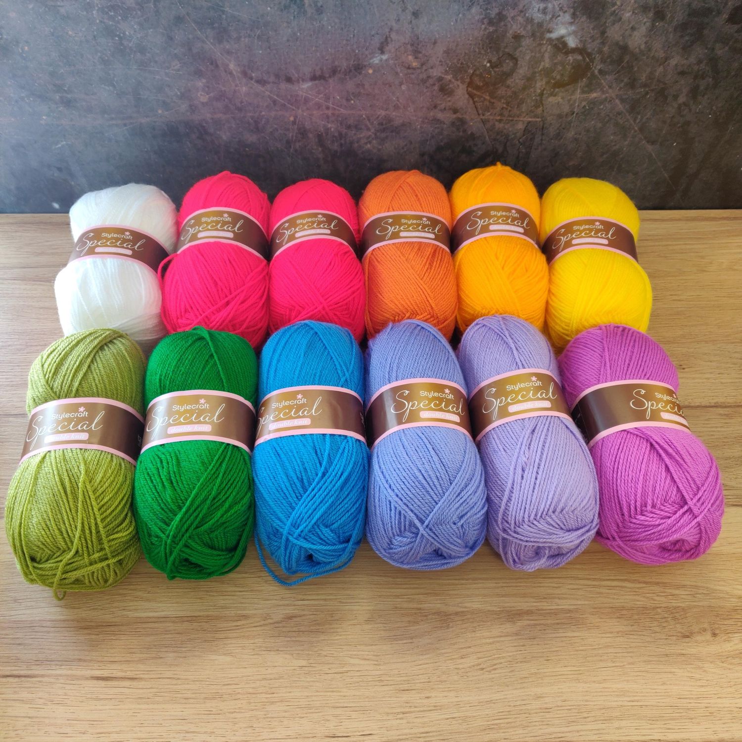 12 balls of yarn in shades of white, red, orange, yellow, green, blue and purple laid out on a wooden surface.