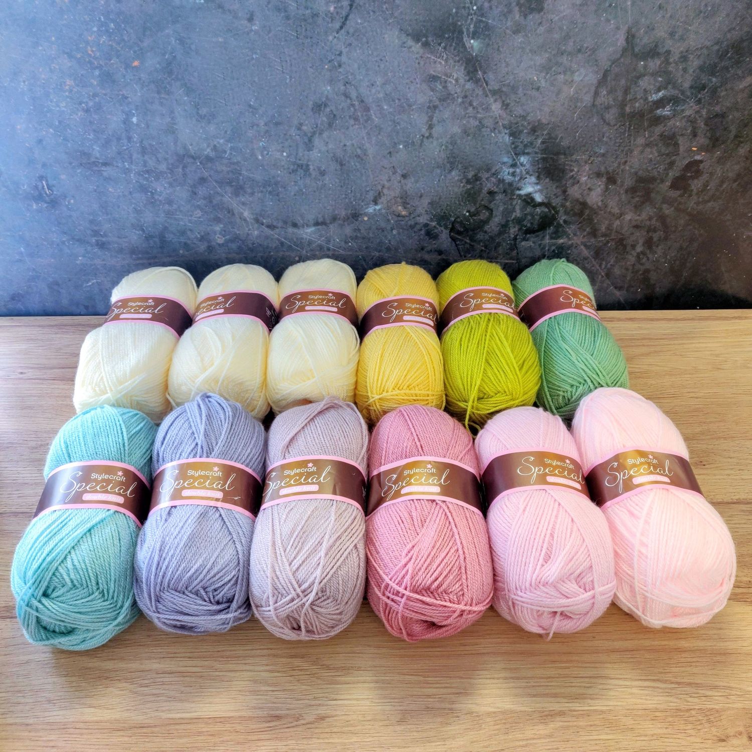12 balls of yarn in shades of cream, green, blue, purple and pink laid out on a wooden surface.