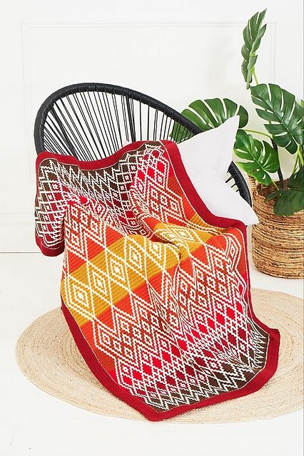 A crocheted blanket in various shades of red and orange, with a cream diamond shaped design.
