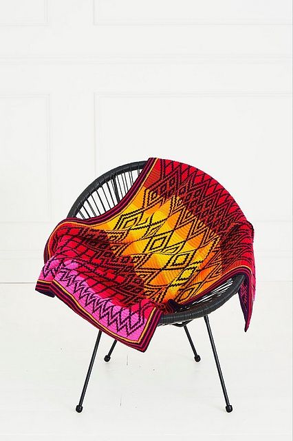 A crocheted blanket in various shades of red, pink, yellow and orange, with a dark purple diamond shaped design.