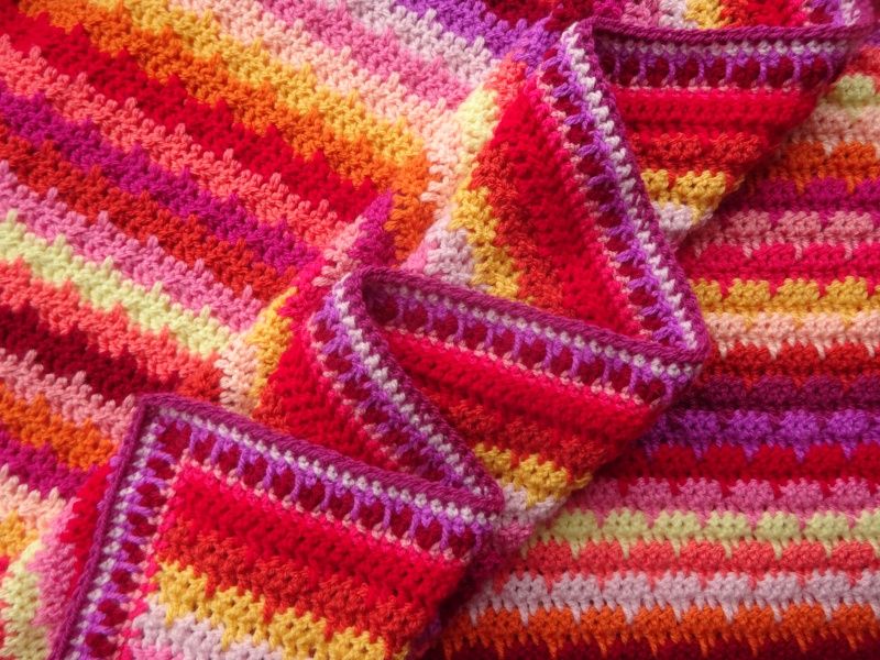 A close up of a multicoloured striped crocheted blanket in shades of pink, purple, red, orange and yellow.
