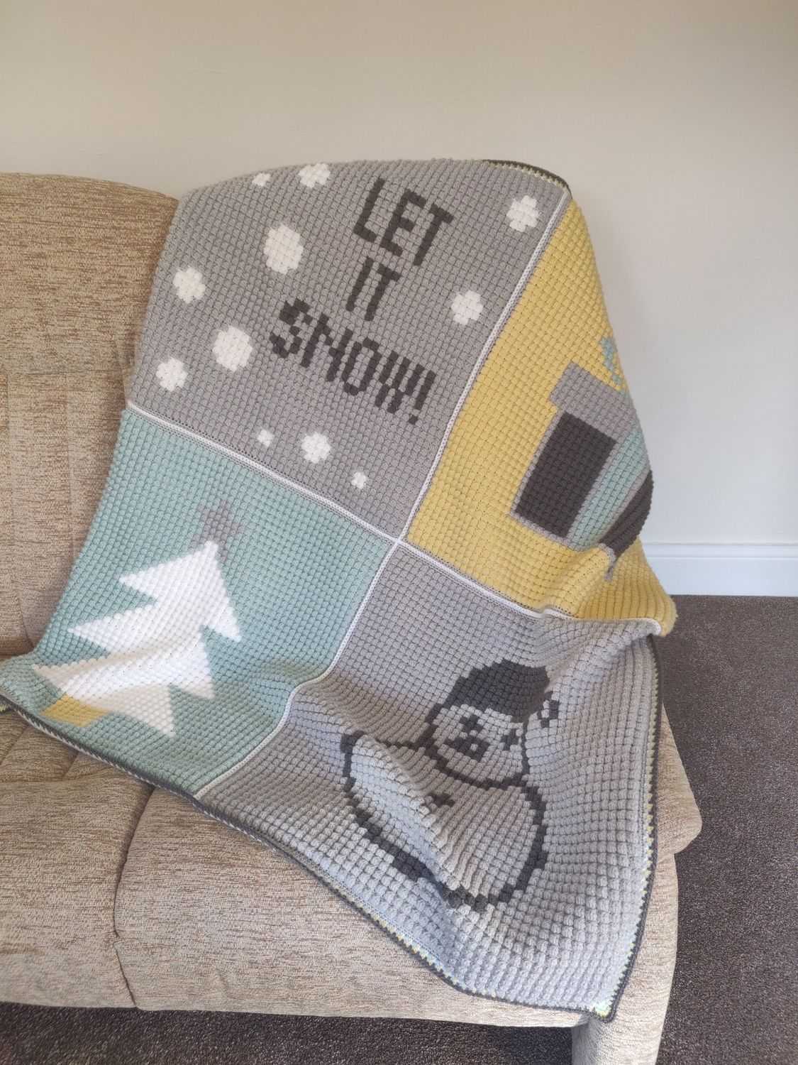 A crocheted blanket made up of 4 large squares, each with a festive design: 'let it snow', a present, a tree and a snowman.