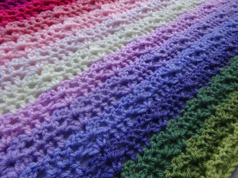 A close up of a striped crochet blanket in shades of pink, purple, cream and green.