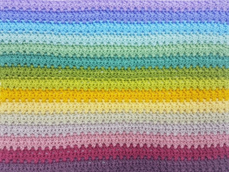 A close up of a striped crochet blanket in shades of purple, blue, green, yellow and pink.