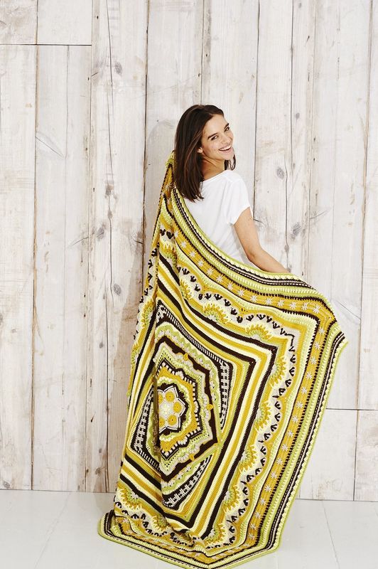 A woman is holding a crocheted blanket in yellow, green, beige and brown.