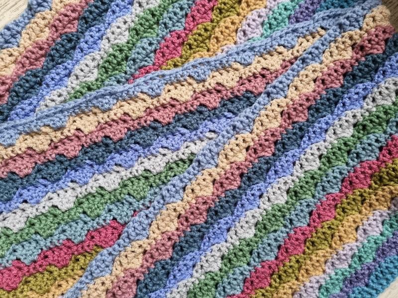 A close up of a crocheted blanket in a wavy stitch in blues, pinks, purples, greens and neutrals.