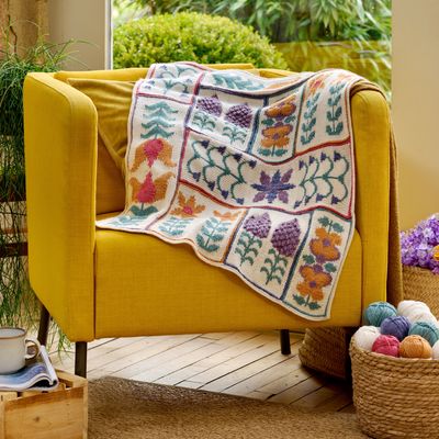 A knitted blanket draped over a yellow chair, featuring yellow and purple flowers and green leaves on a cream background.