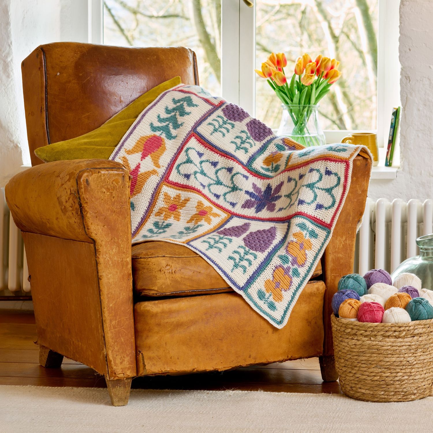 A crocheted blanket draped over a chair, featuring yellow and purple flowers and green leaves on a cream background.