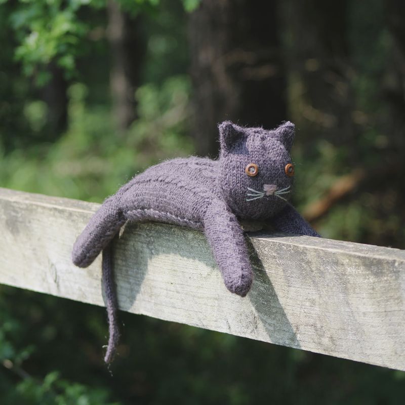 A dark grey knitted cat, with cable stitches on it's body.