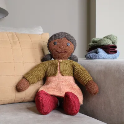 A knitted doll toy with brown skin, black hair, a peach dress, red shoes and a green cardigan.