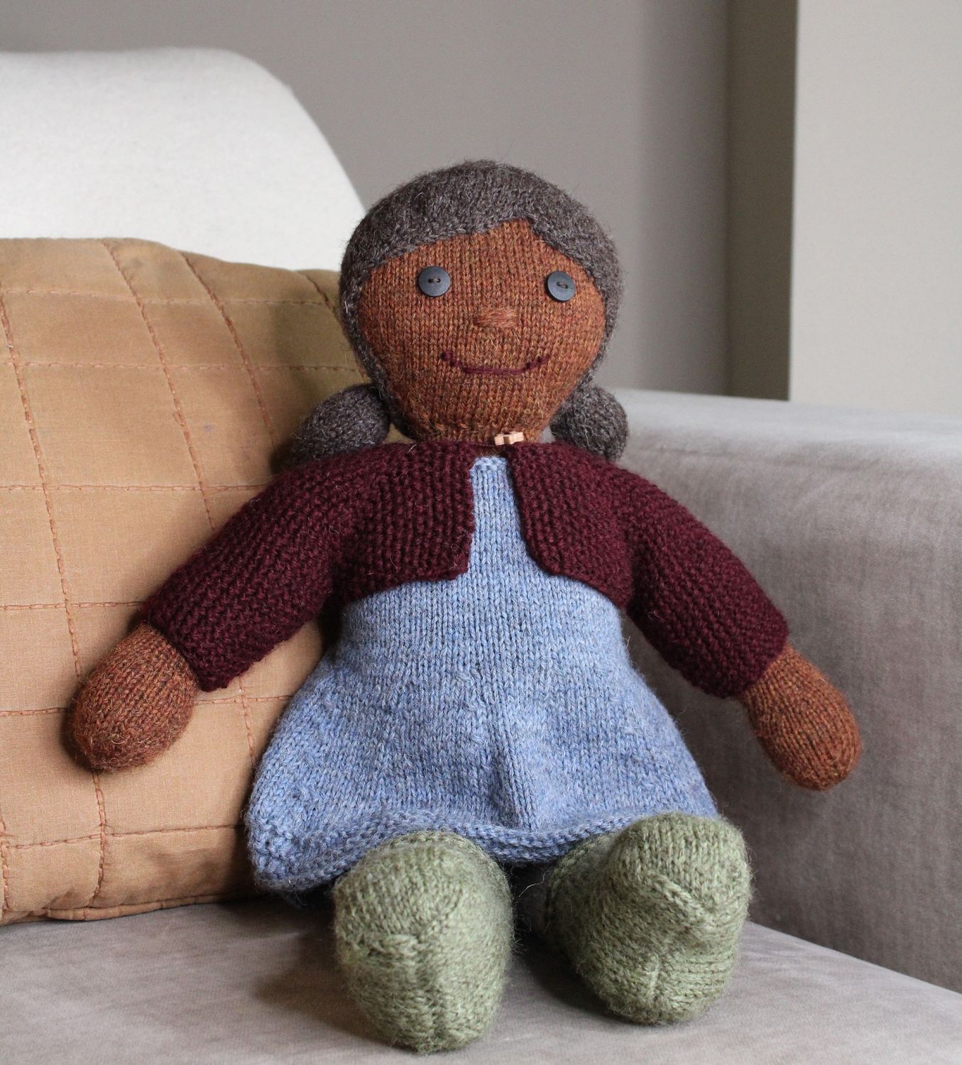 A knitted doll toy with brown skin, black hair, a blue dress, green shoes and a burgundy red cardigan.