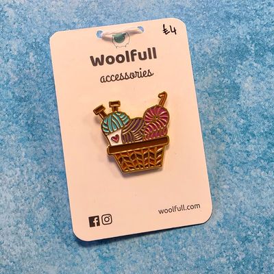 A pin badge of a basket containing balls of yarn.