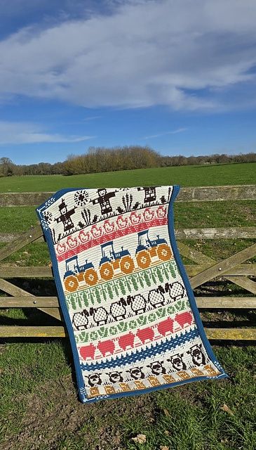 A crocheted blanket in cream, blue, pink, green and brown with colourwork scarecrows, tractors, sheep, pigs and chickens.