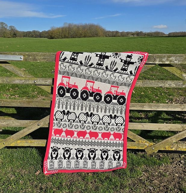 A crocheted blanket in silver, grey, pink and black with colourwork scarecrows, tractors, sheep, pigs and chickens.