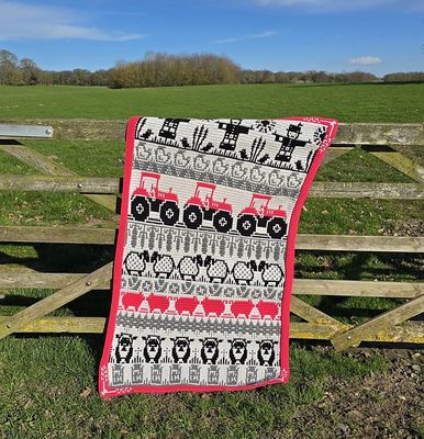 A crocheted blanket in silver, grey, pink and black with colourwork scarecrows, tractors, sheep, pigs and chickens.