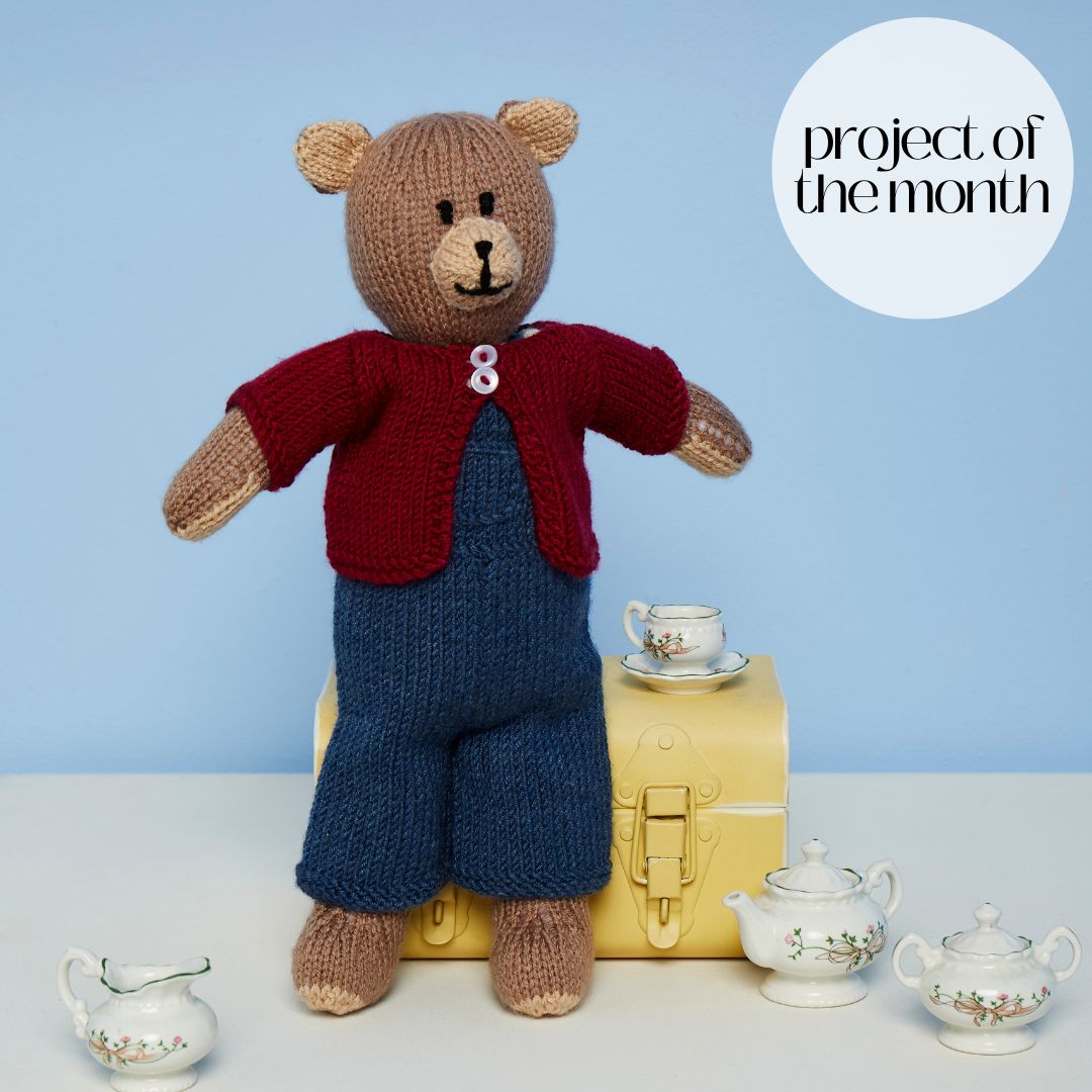 A brown knitted bear toy wearing blue knitted dungarees and a red knitted cardigan.