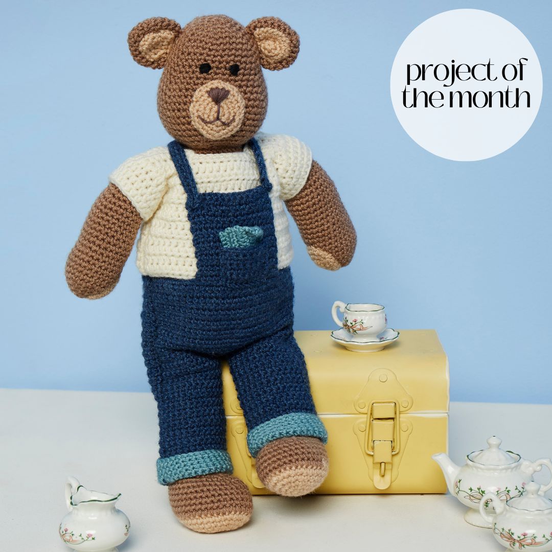 A brown crocheted bear wearing blue crocheted dungarees and a cream crocheted top.