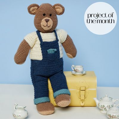 A brown crocheted bear wearing blue crocheted dungarees and a cream crocheted top.