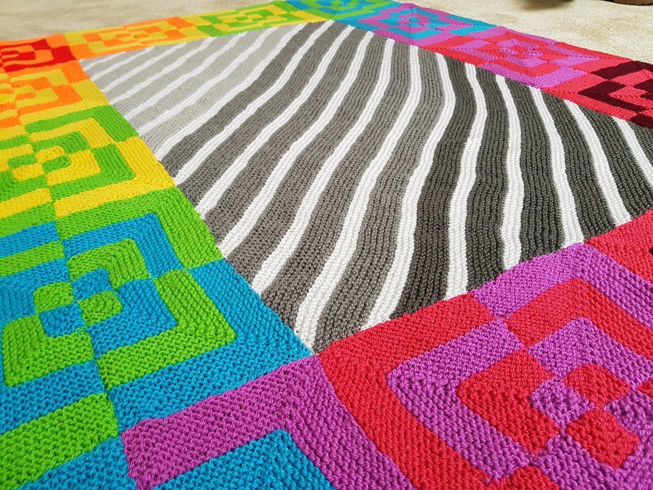 A knitted blanket, with a white and grey striped centre surrounded by brightly coloured squares.