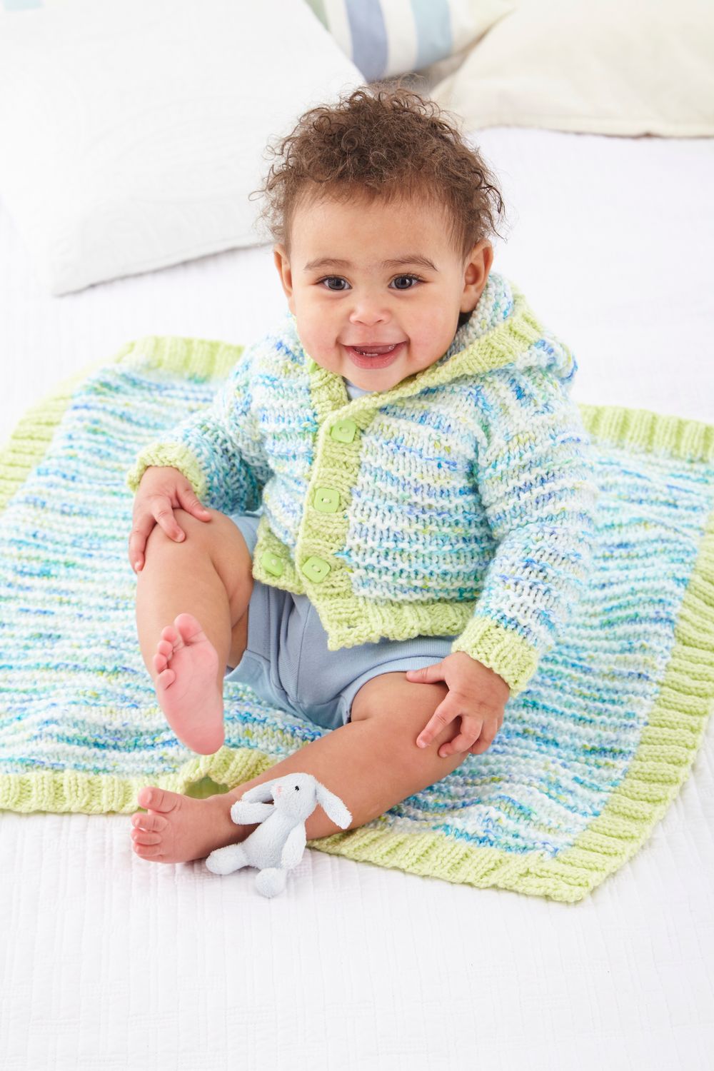 A baby is sitting on a blanket and wearing a knitted cardigan, both in a white, green and blue variegated yarn.