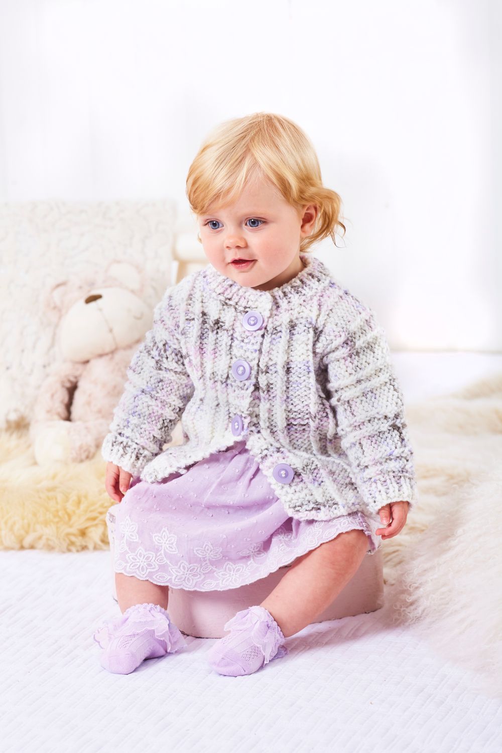 A baby is wearing a knitted cardigan in a white, purple and grey variegated yarn.