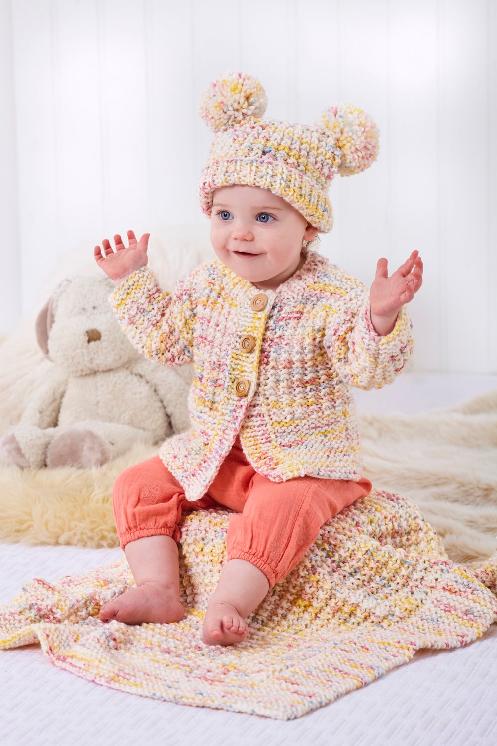 A baby is wearing a knitted cardigan and hat in a white, yellow and pink variegated yarn.