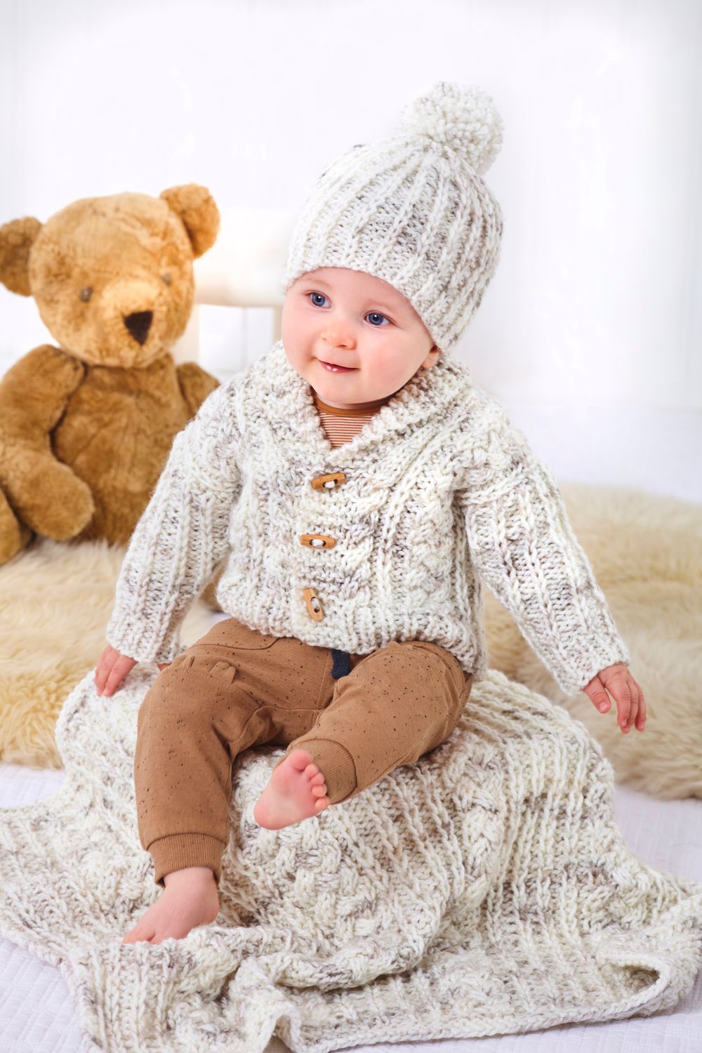 A baby is wearing a knitted cardigan and hat in a cream and brown variegated yarn.