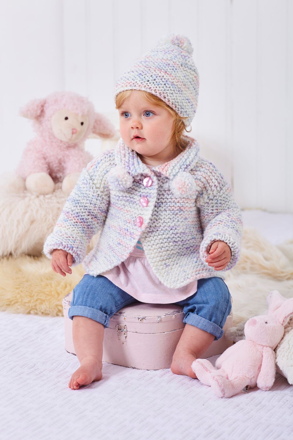 A baby is wearing a knitted cardigan and hat in a pink, purple and white variegated yarn.