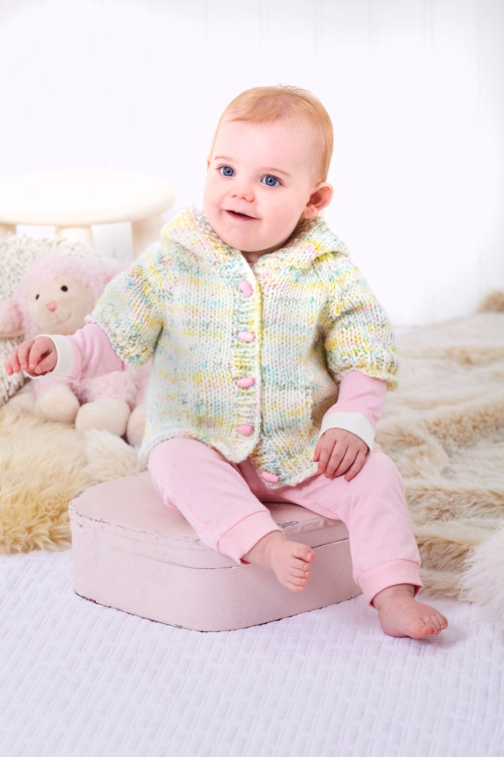 A baby is wearing a knitted cardigan and hat in a white, yellow and blue variegated yarn.