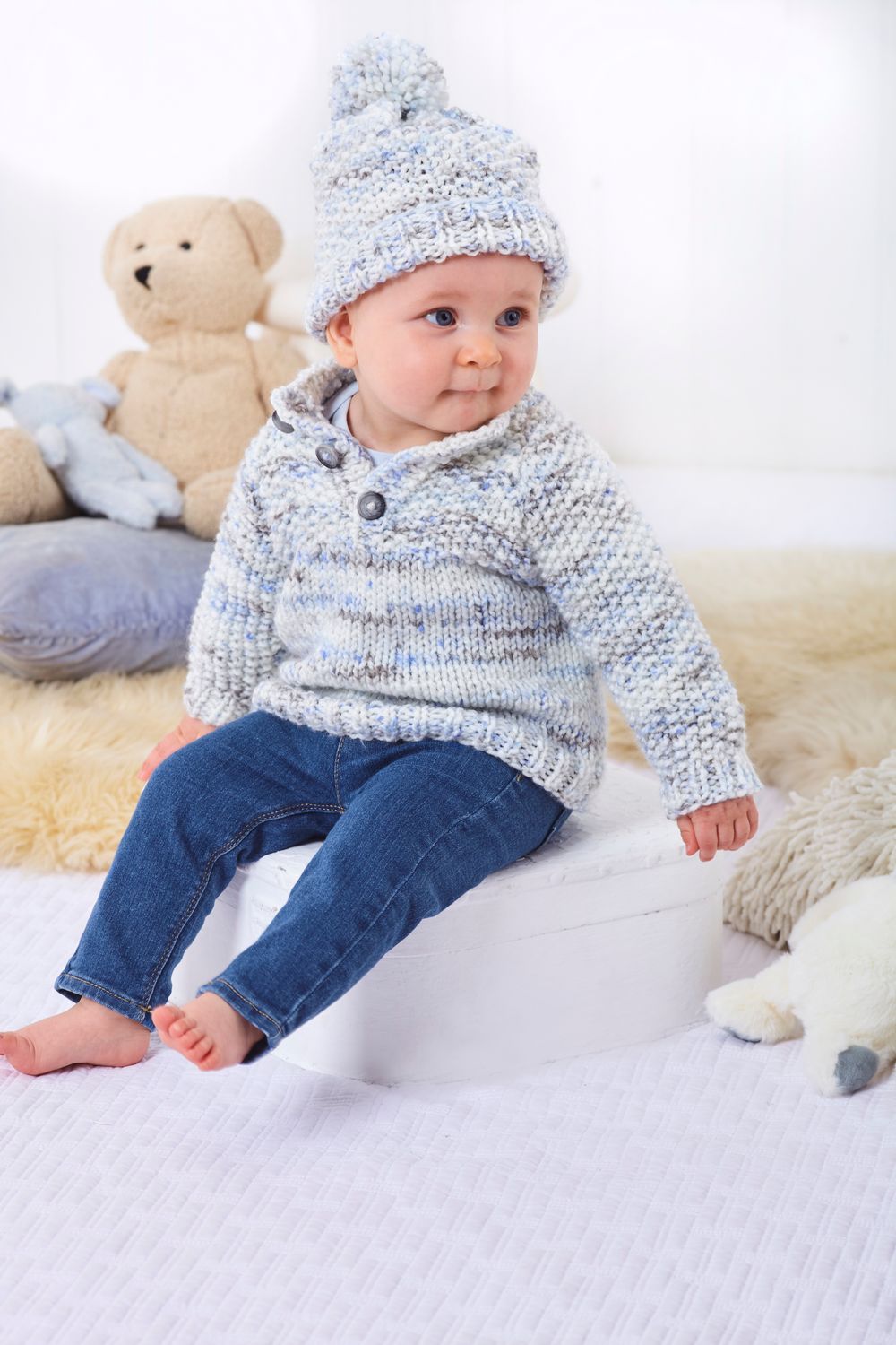 A baby is wearing a knitted sweater and hat in a blue, grey and white variegated yarn.