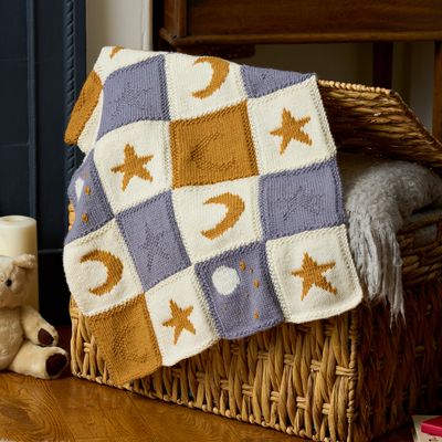 A knitted blanket made up of cream, blue and gold squares with moon and star motifs.