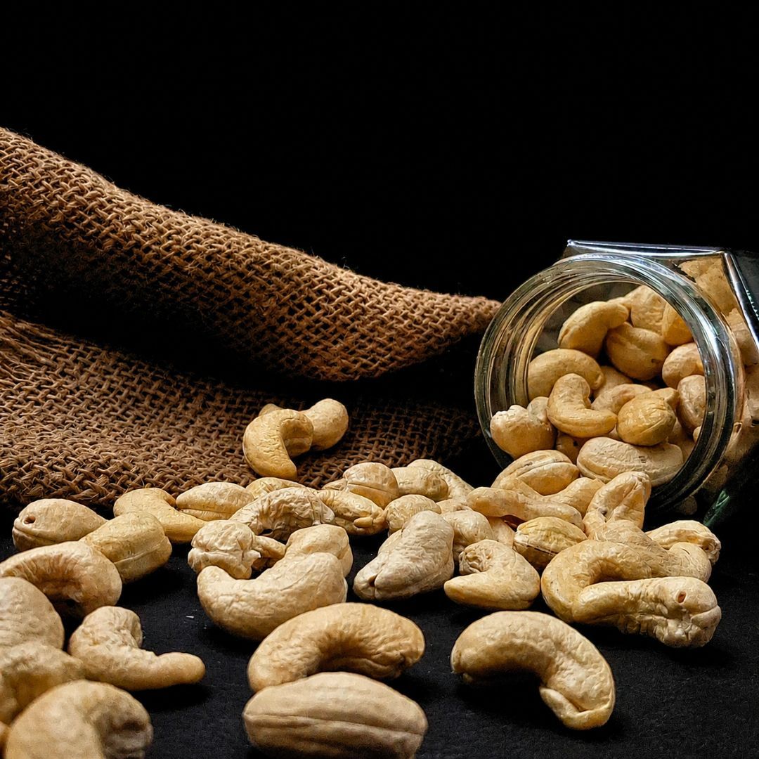 Organic Konkan W210 cashews scattered beside a glass jar on rustic burlap, showcasing their large size and natural texture.