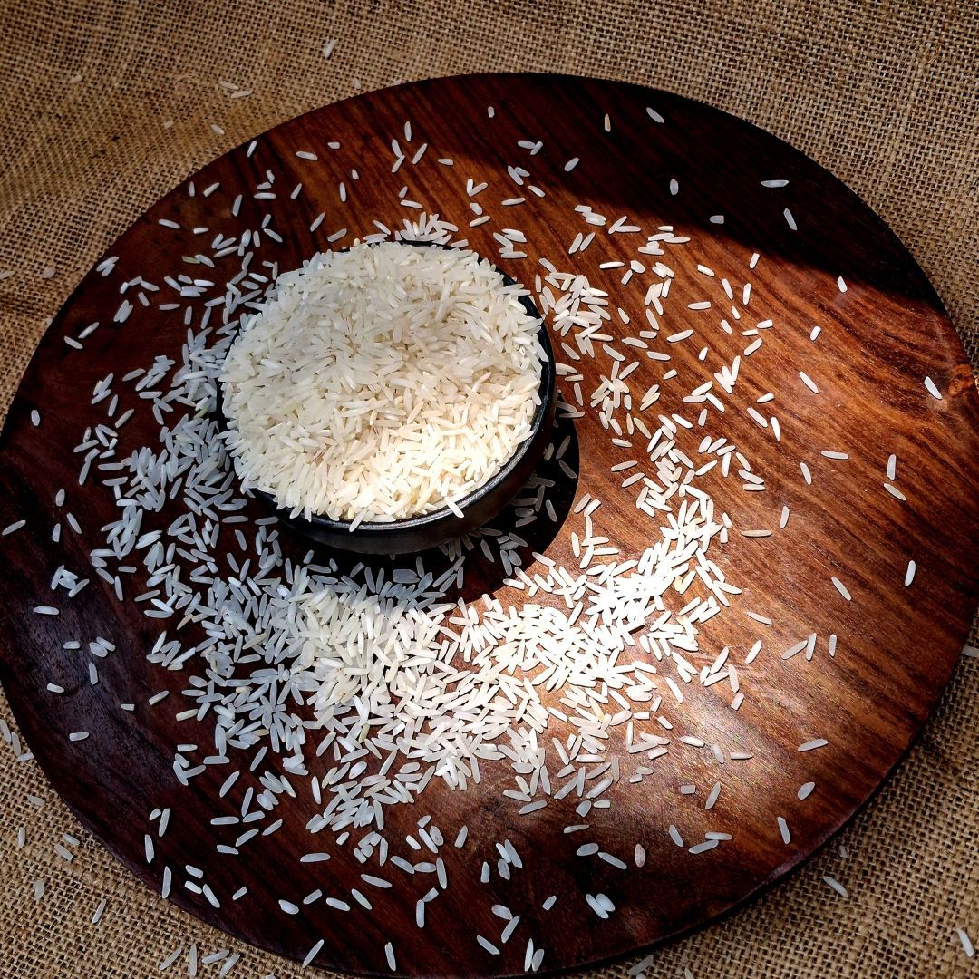 Organic Tarawadi Basmati White Rice in a black bowl, surrounded by loose grains on a wooden platter highlighting long grains.