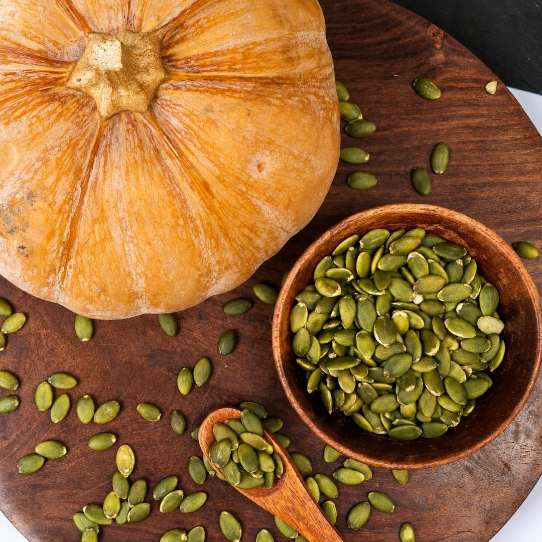 A rustic harvest scene featuring green pumpkin seeds, a whole pumpkin and scattered seeds across a wooden surface.