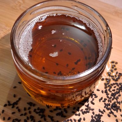 Organic black sesame oil in a glass jar, with black sesame seeds scattered around, highlighting its rich amber color. earthy.