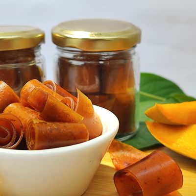 Organic Mango Roll Aam Papad in a bowl with rolled pieces, jars and fresh mango slices showcasing its fruity texture.
