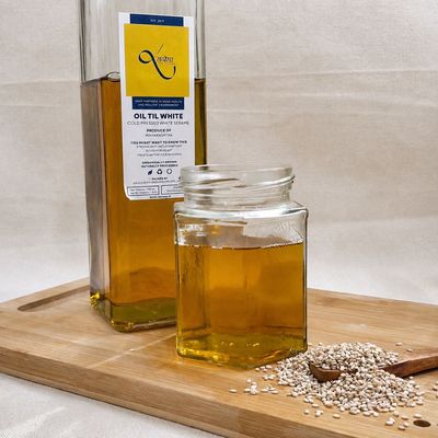 Organic white sesame oil in glass bottles, cold-pressed for pure flavor, with white sesame seeds displayed beside it on wood.