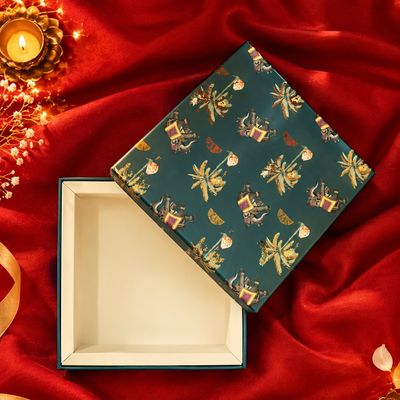 Small festive sweet box with a patterned green lid, ivory interior and red satin backdrop, beside a glowing Diwali diya lamp.
