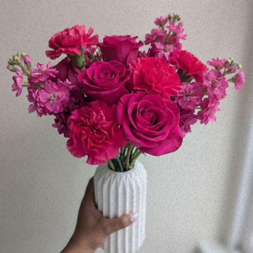 Bright pink and fuchsia floral arrangement in a white fluted ceramic vase, featuring carnations, stock, and roses.