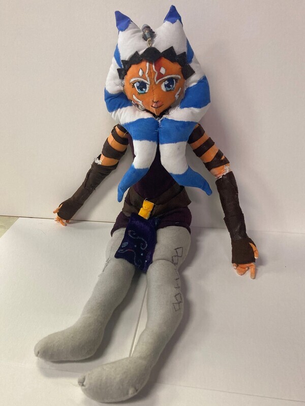 Ahsoka Tano is a 20&quot; doll of the Star Wars character Ahoska. Ahoska has a white and blue striped head tails or lekku that extend over her chest. Her lekku are held back by a brown headband with three gold gems on it. Her skin is an orange brown with white marks over her eyebrows and blue eyes that continue down her cheeks. On her arms are three bands and her long fingerless gloves. Ahsoka wears a brown shirt with a brown gold buckle belt. She has a blue skirt over grey leggings/ boots. 