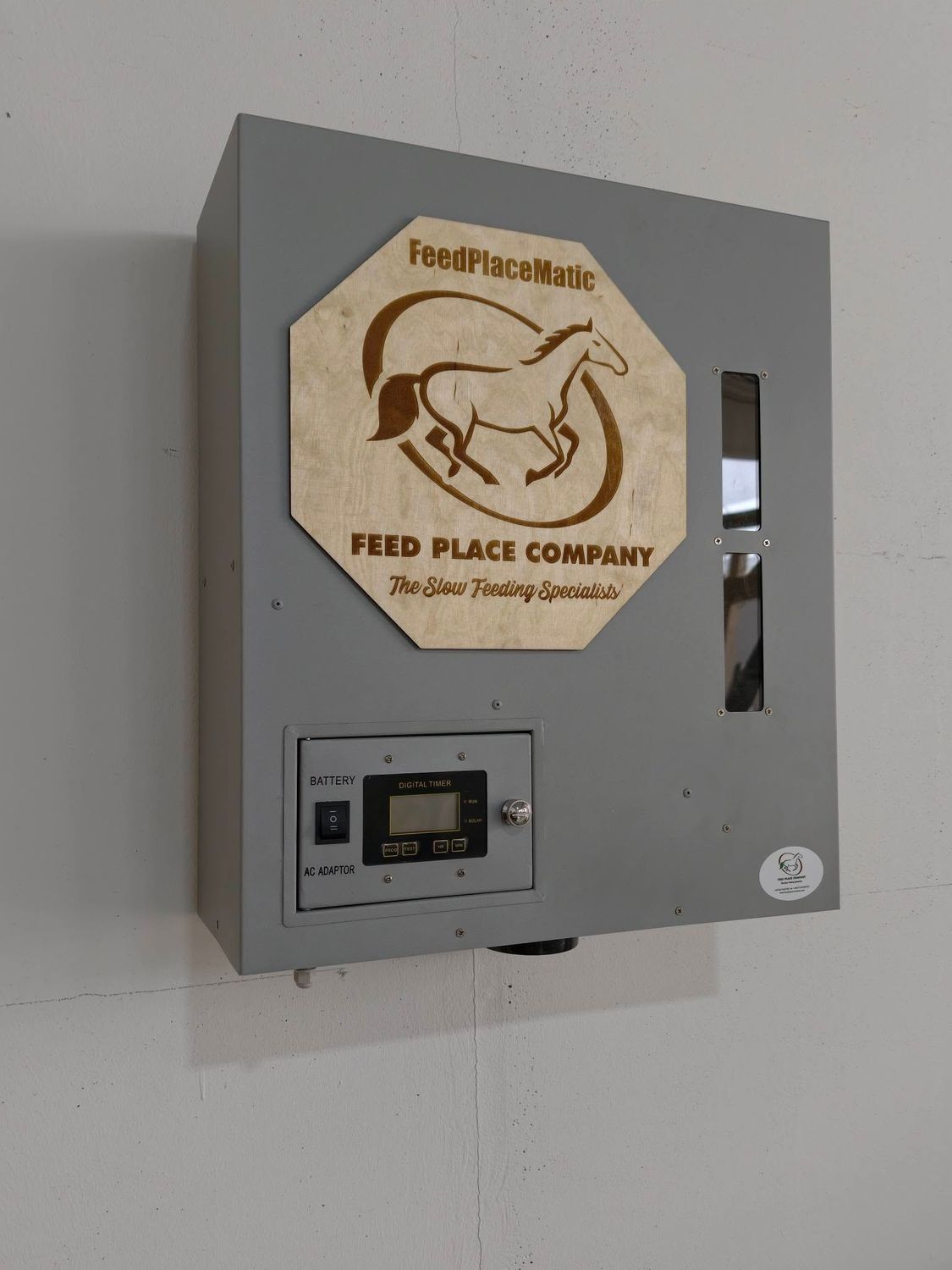 FeedPlaceMatic horse feeder by FEED PLACE COMPANY, gray metal box with digital display and battery backup, mounted on wall.