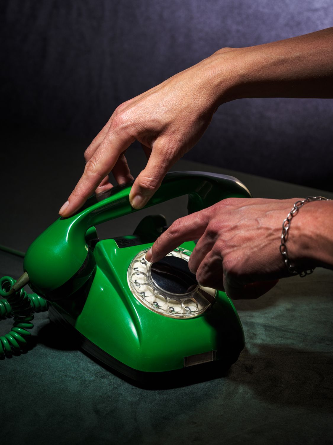 fotografia kolekcjonerska przedstawiająca zielony aparat telefoniczny retro z lat 60-tych oraz dłonie - jedna podnosi słuchawkę a druga wybiera numer. kolory tła to szara zieleń i ciemny granat. całość sfotografowana w enigmatyczny sposób.