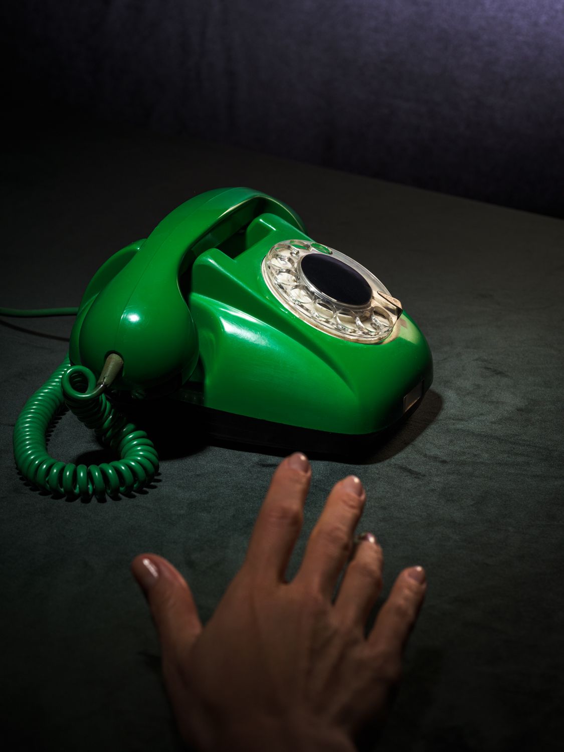 fotografia kolekcjonerska przedstawiająca zielony aparat telefoniczny retro z lat 60-tych oraz rękę wyciągniętą w kierunku słuchawki. kolory tła to szara zieleń i ciemny granat. całość sfotografowana w enigmatyczny sposób.