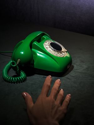 fotografia kolekcjonerska przedstawiająca zielony aparat telefoniczny retro z lat 60-tych oraz rękę wyciągniętą w kierunku słuchawki. kolory tła to szara zieleń i ciemny granat. całość sfotografowana w enigmatyczny sposób.