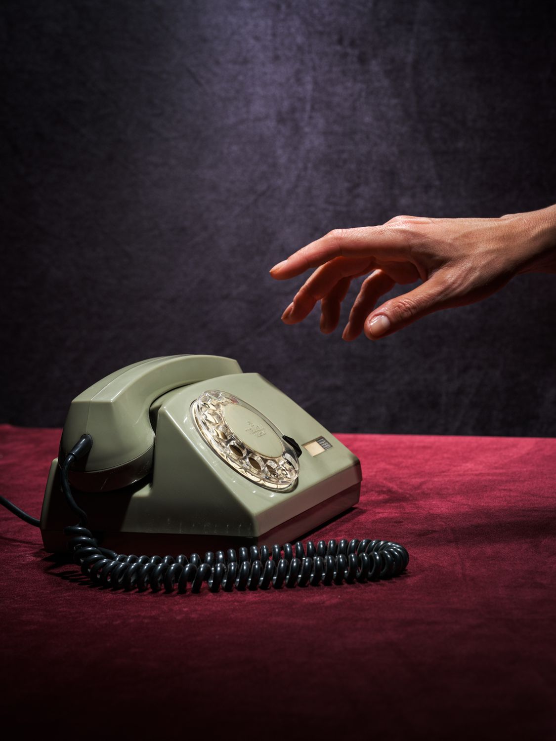 fotografia kolekcjonerska przedstawiająca szary aparat telefoniczny retro z lat 70-tych oraz rękę wyciągniętą w kierunku słuchawki. kolory tła to czerwień i ciemny granat. całość sfotografowana w enigmatyczny sposób.