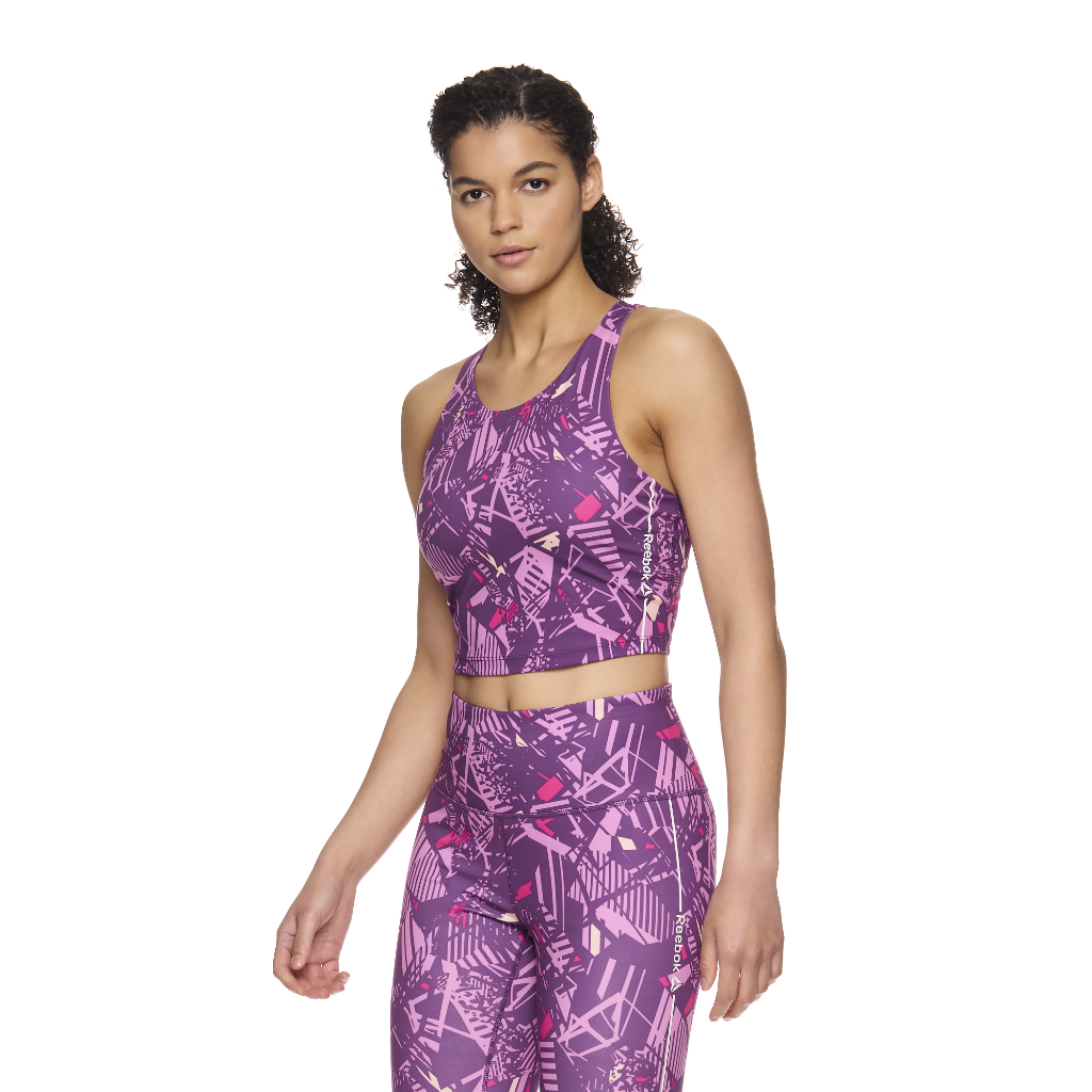 A woman wearing a Reebok Women&#39;s Cropped Tank Bra in Sunset Purple, featuring a bold abstract pattern. The cropped tank has a racerback design and a snug, flattering fit. The fabric is stretchy and breathable, suitable for workouts or casual wear. The model is showcasing the front, side, and back views of the tank bra. Size XS, brand new with tags.