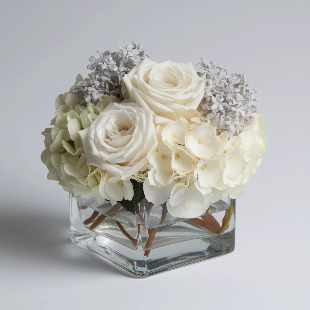 An elegant floral arrangement of white hydrangeas, roses, and grey carnations in a clear glass cube vase 